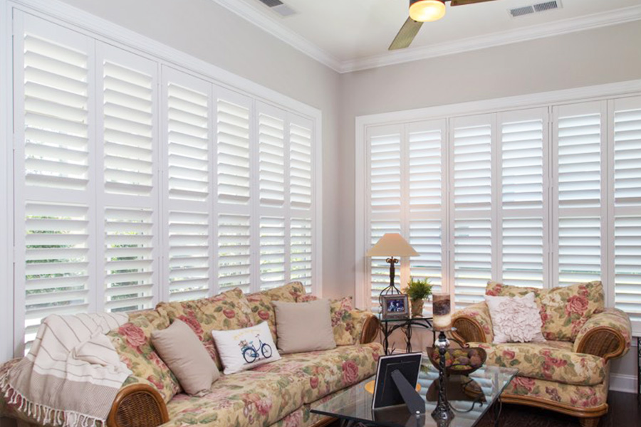 Use interior shutters to manage light levels and temperature changes in your sunroom to keep it comfortable. White Polywood shutters behind floral couches in a sunroom